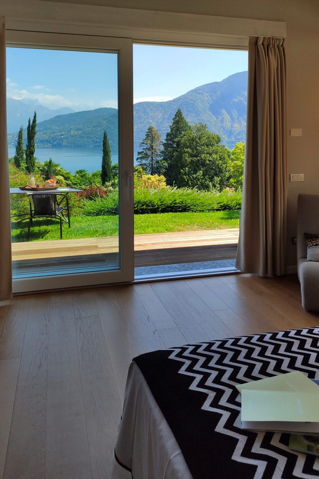 Interior view of a modern room with large glass doors opening to a lush garden and scenic lake view, featuring wooden flooring and natural light. | Only on SopranoVillas