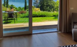 Interior view of a modern room with large glass doors opening to a lush garden and scenic lake view, featuring wooden flooring and natural light.