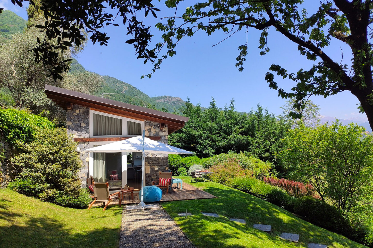 Charming modern cottage surrounded by lush greenery, featuring a patio with outdoor seating and an umbrella, set against a backdrop of mountains and blue sky. Ideal for a tranquil getaway or nature retreat. | Only on SopranoVillas