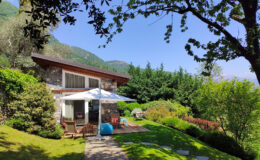 Charming modern cottage surrounded by lush greenery, featuring a patio with outdoor seating and an umbrella, set against a backdrop of mountains and blue sky. Ideal for a tranquil getaway or nature retreat.