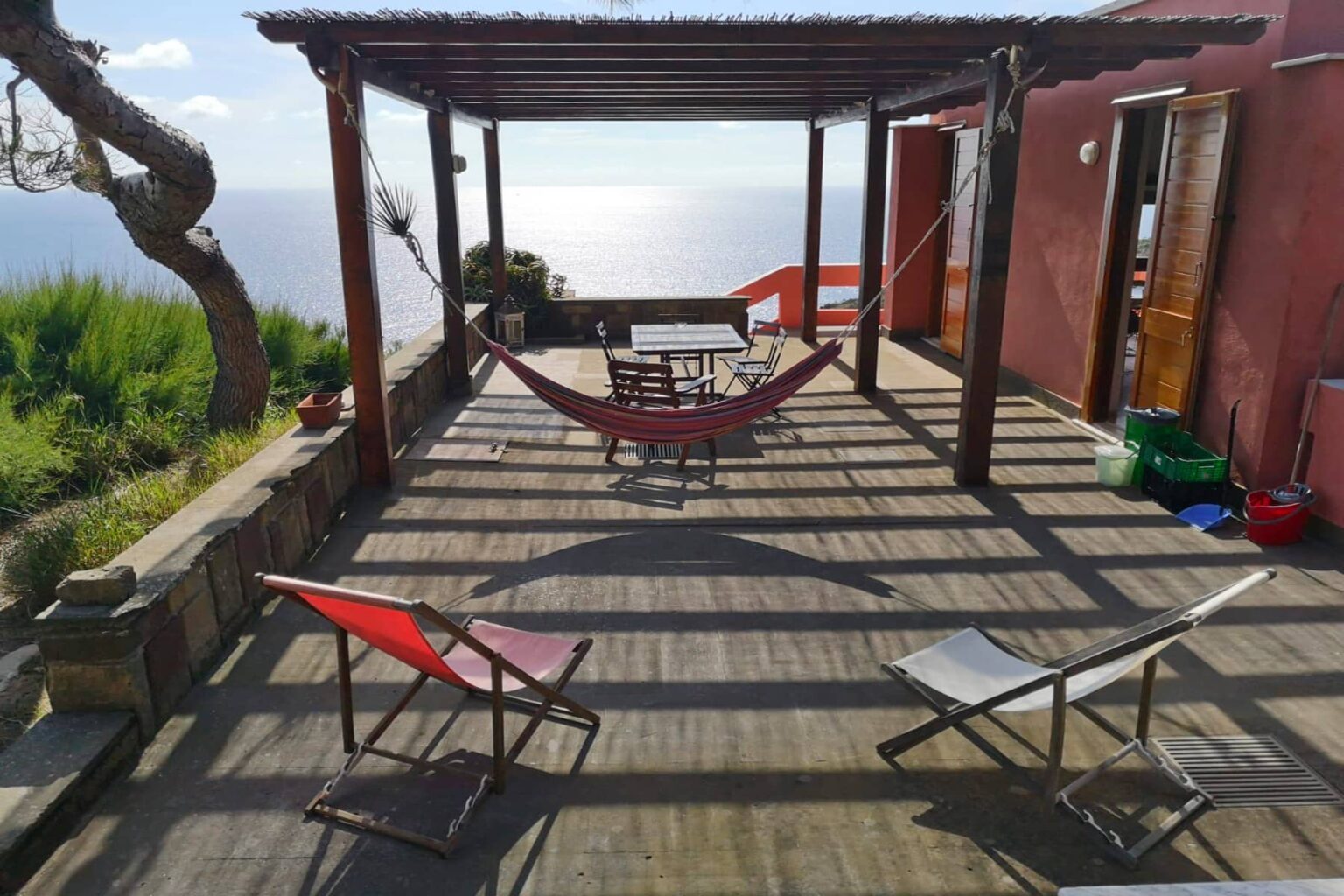 Cozy outdoor patio with a hammock and lounge chairs overlooking the ocean, featuring wooden flooring and a rustic building in the background. Ideal for relaxation and enjoying scenic views. | Only on SopranoVillas