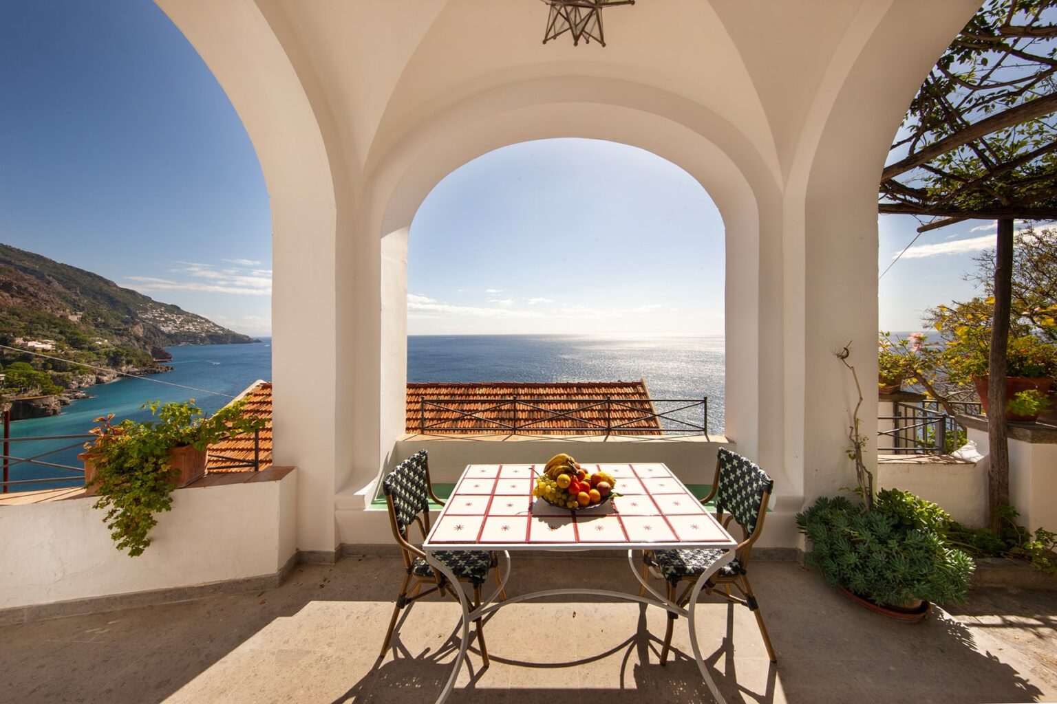 Cozy outdoor dining area with a table set for breakfast, overlooking a serene ocean view, framed by an elegant archway and lush greenery. | Only on SopranoVillas