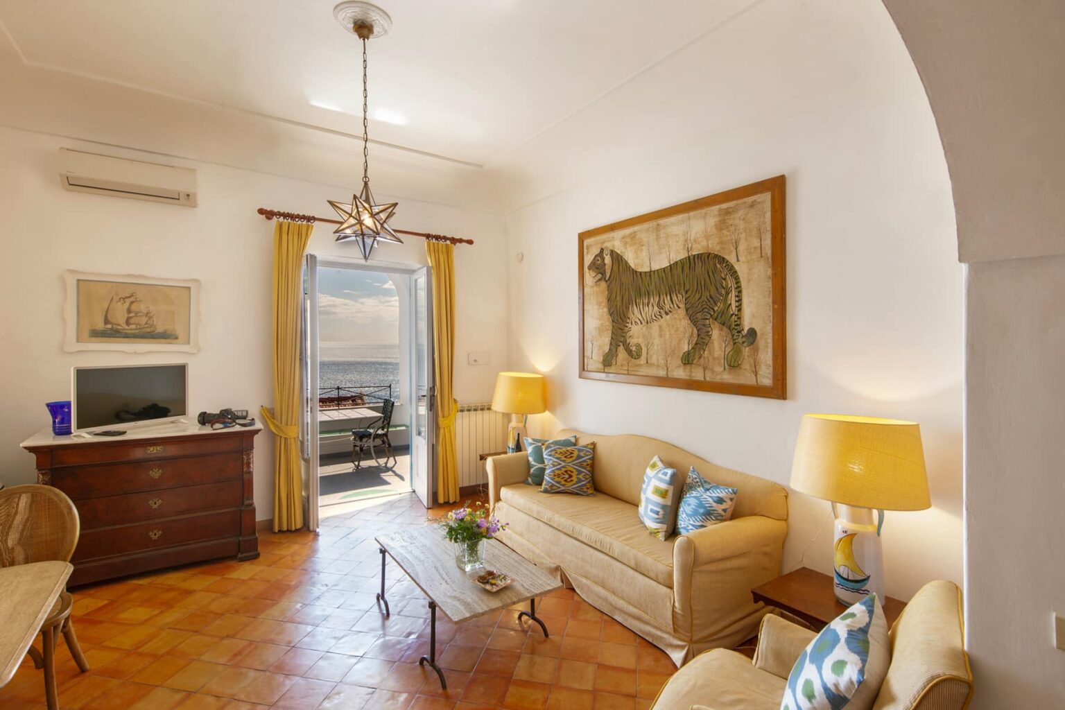 Cozy living room with beige sofas and decorative pillows, a large lion painting on the wall, wooden furniture, and natural light from an open door leading to a balcony with a view. | Only on SopranoVillas