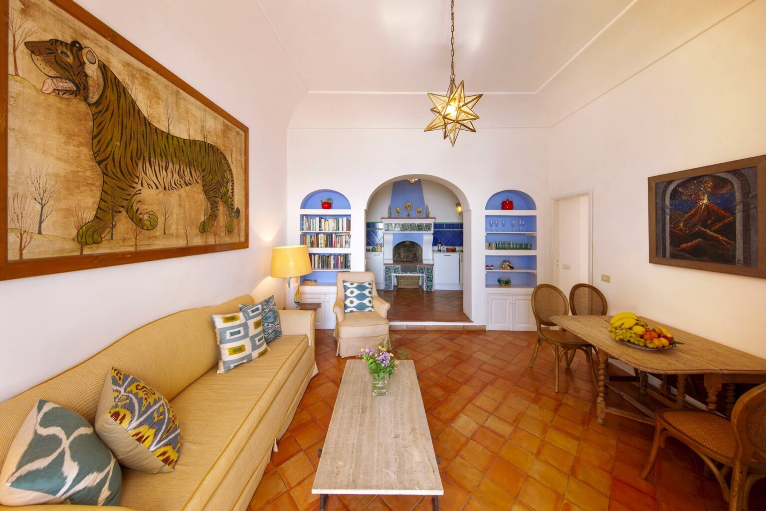 Spacious living room featuring a decorative tiger mural, stylish beige sofa with colorful cushions, a wooden dining table, and a bright chandelier, creating a warm and inviting atmosphere. | Only on SopranoVillas