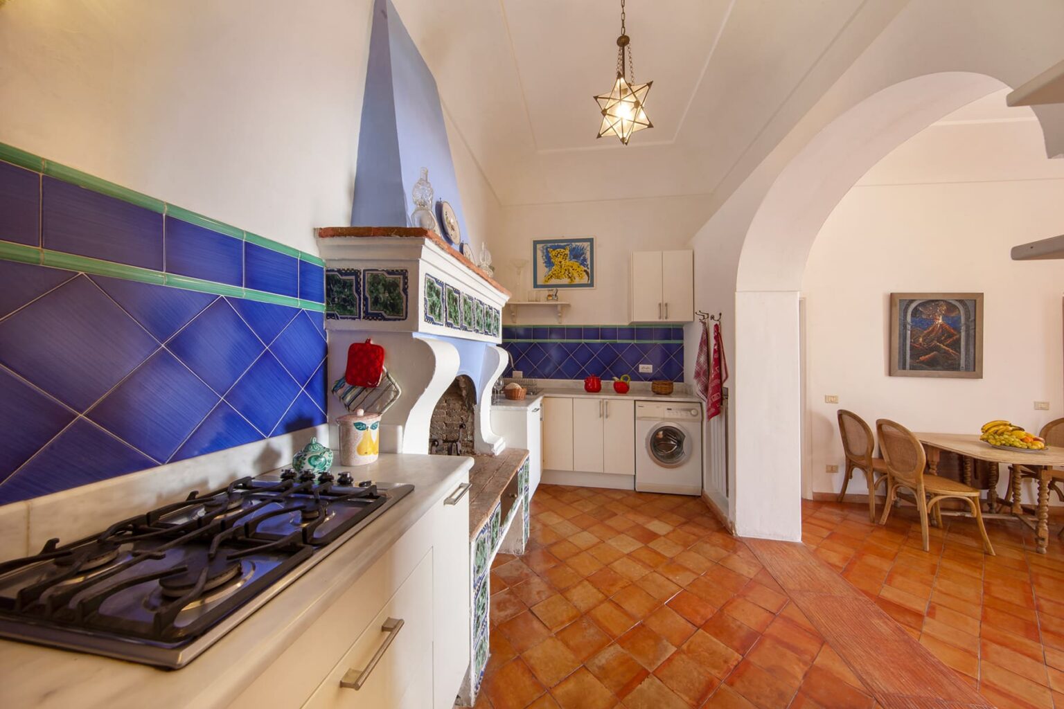 Bright and spacious kitchen featuring a traditional design with blue tiled backsplash, a gas stove, and terracotta floor tiles. The kitchen includes a dining area with wooden chairs and a decorative chandelier, creating a warm and inviting atmosphere. | Only on SopranoVillas