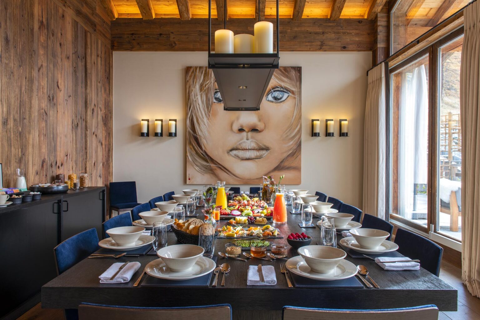 Elegant dining room featuring a long table set for a meal, adorned with plates and colorful food arrangements. A striking wall art piece of a woman's face adds a contemporary touch, while wooden accents and large windows create a warm, inviting atmosphere. Ideal for showcasing luxury dining experiences. | Only on SopranoVillas