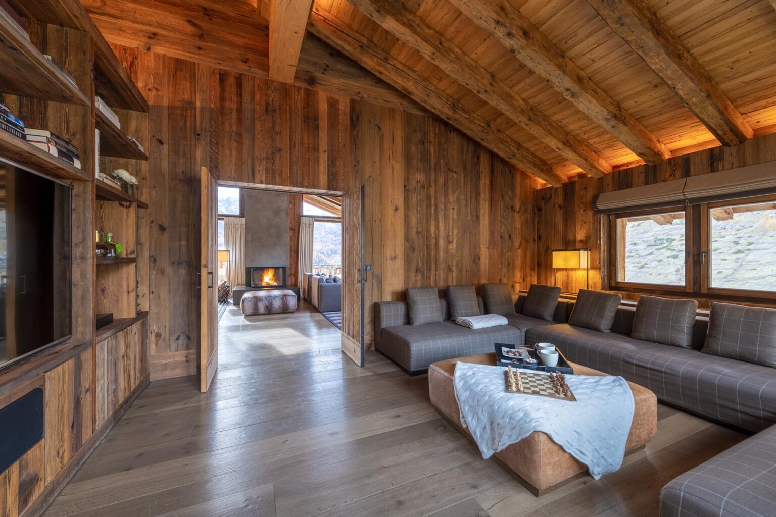 Modern rustic living room with wooden walls, a cozy sectional sofa, and a warm ambiance, featuring large windows that provide natural light and a view of the outdoors. | Only on SopranoVillas