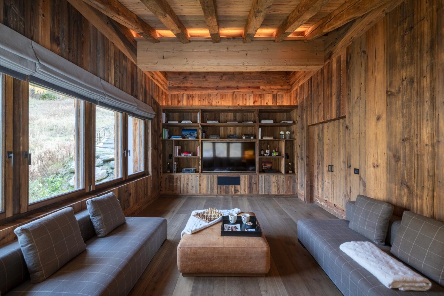Modern rustic living room featuring wooden walls, large windows, and comfortable seating arrangements. The space includes a cozy coffee table and a stylish shelving unit, creating a warm and inviting atmosphere. | Only on SopranoVillas