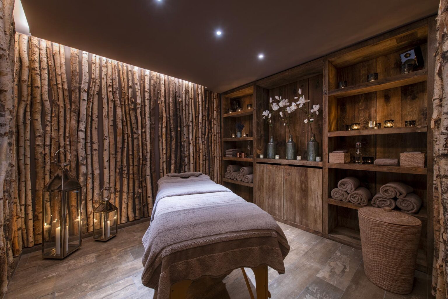 Spa treatment room featuring a massage table, wooden decor, ambient lighting, and shelves with wellness products. The walls are adorned with natural wood, creating a calming atmosphere for relaxation and rejuvenation. | Only on SopranoVillas