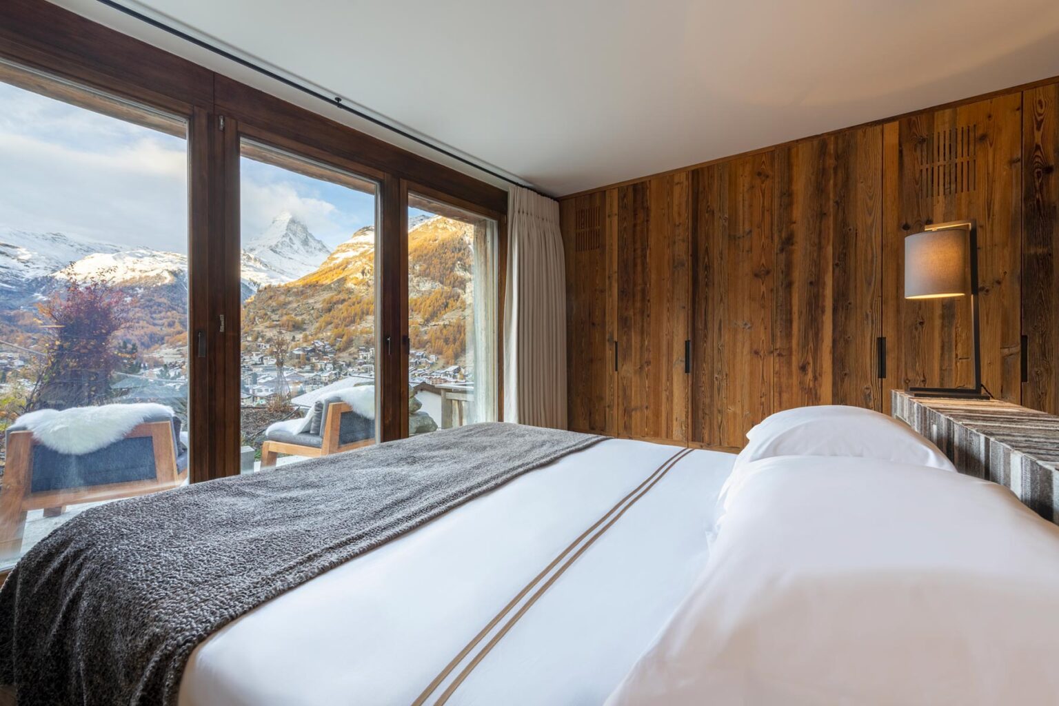 Cozy bedroom with wooden accents and large windows showcasing a mountain view, featuring a neatly made bed with white linens and a gray throw blanket. | Only on SopranoVillas
