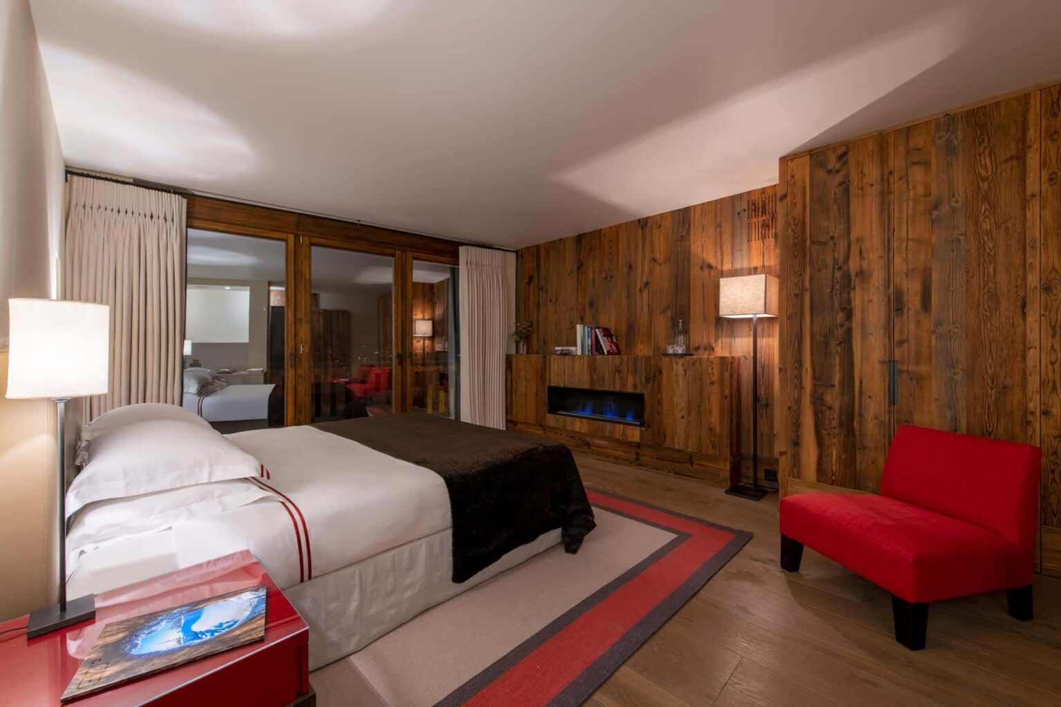Cozy hotel room featuring a wooden accent wall, a comfortable queen-sized bed with white linens, a bright red accent chair, and soft lighting for a warm ambiance. | Only on SopranoVillas