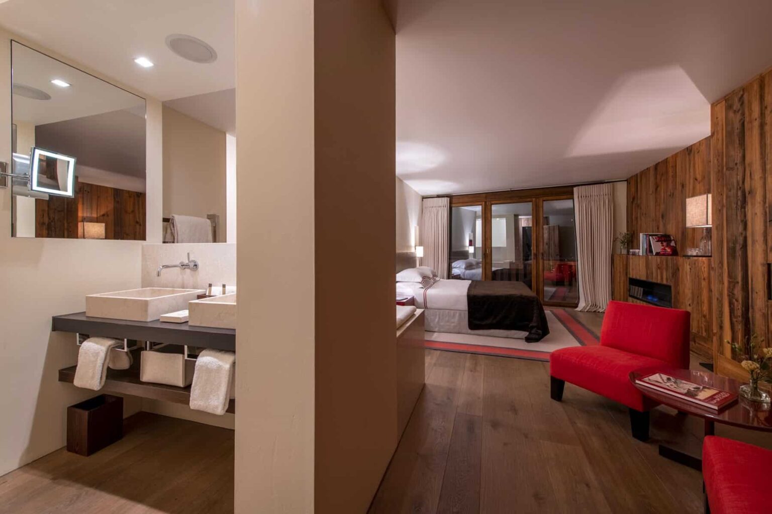 Luxurious hotel room interior featuring a modern bathroom with a stylish sink, a cozy bedroom area with a black bed, and a red accent chair, illuminated by soft lighting. | Only on SopranoVillas