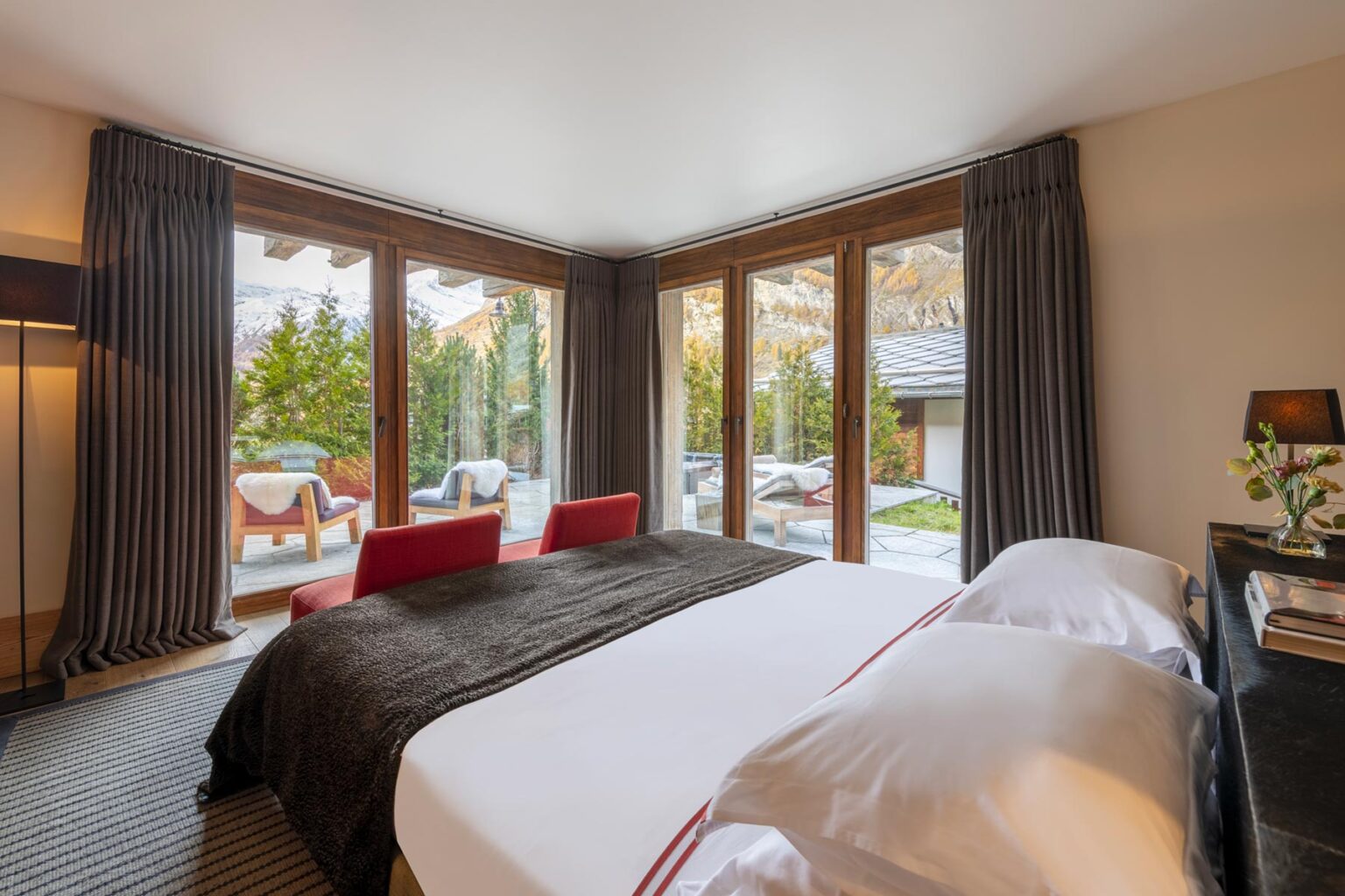 Modern bedroom interior with large windows showcasing a serene outdoor view, featuring a king-size bed with plush bedding, stylish red chairs, and elegant decor, ideal for a relaxing getaway. | Only on SopranoVillas