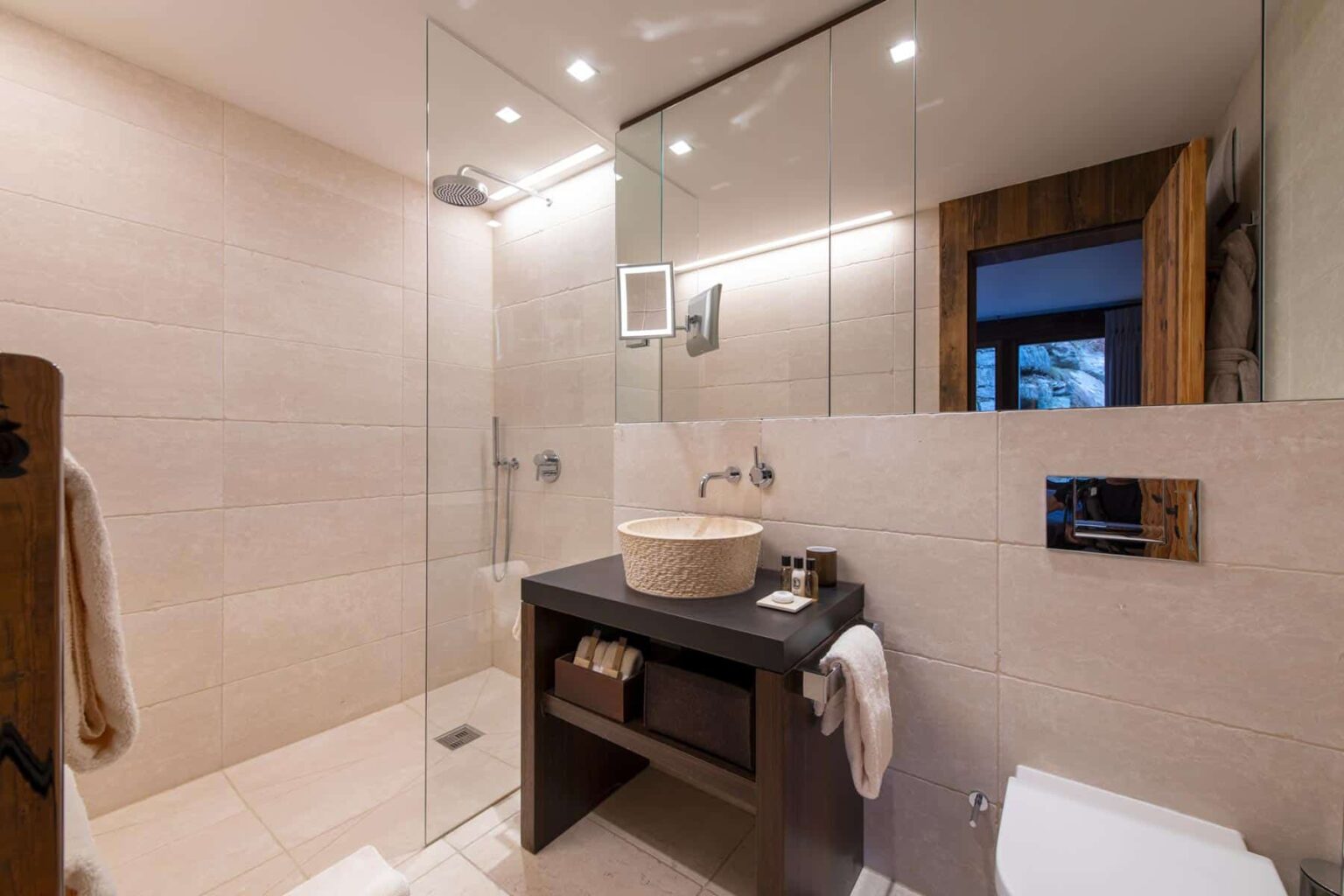 Modern bathroom design featuring a glass shower enclosure, a stylish vessel sink on a dark vanity, and neutral stone tiles. The space is well-lit with recessed lighting, showcasing a contemporary aesthetic. | Only on SopranoVillas
