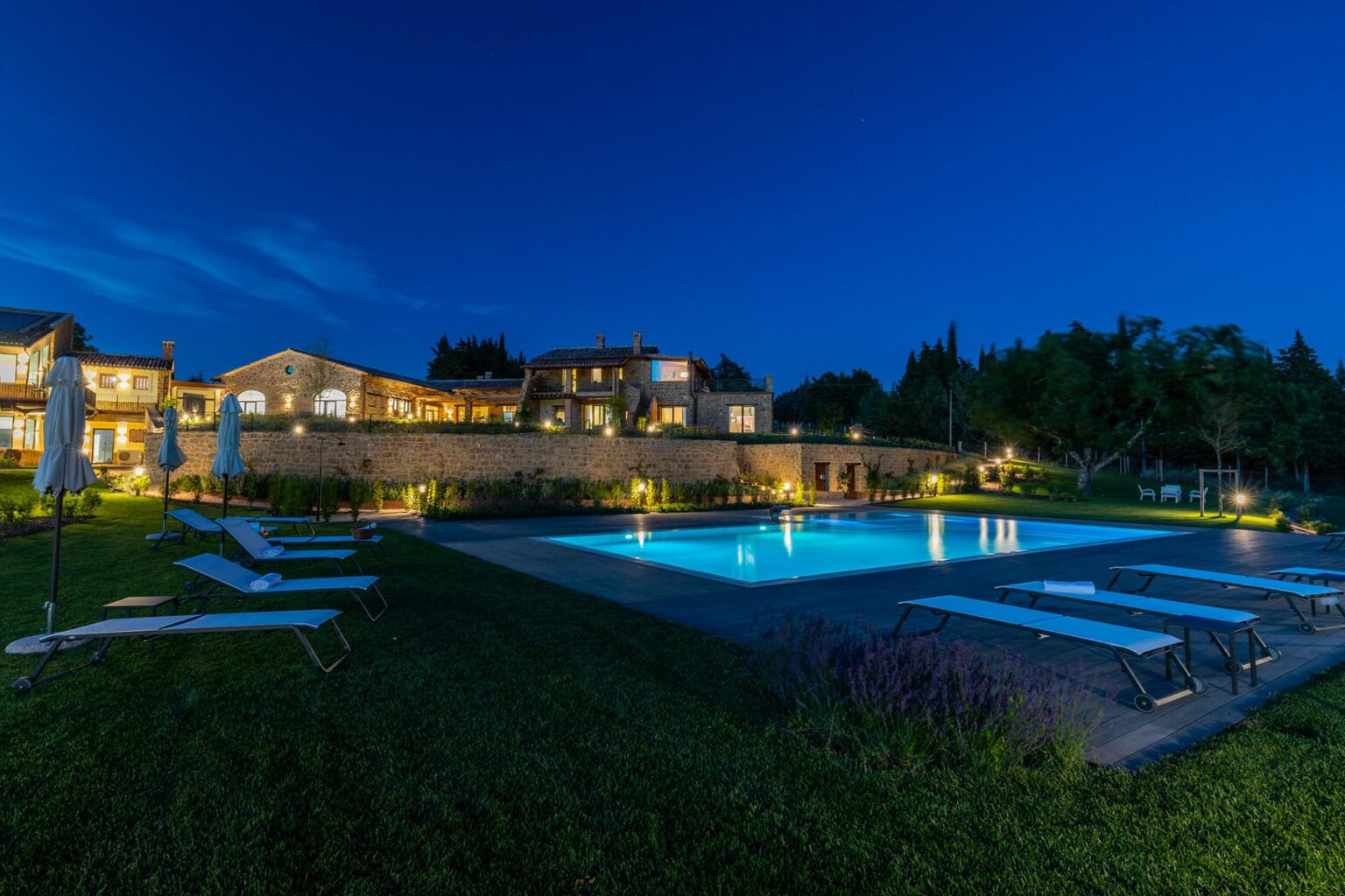 Luxurious villa at dusk featuring a well-lit swimming pool surrounded by lounge chairs and landscaped gardens. | Only on SopranoVillas