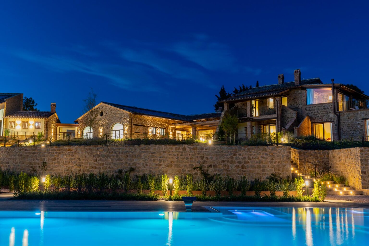 Luxurious stone villa illuminated at night, featuring a sparkling pool in the foreground and beautifully landscaped gardens, showcasing upscale architecture and serene ambiance. | Only on SopranoVillas