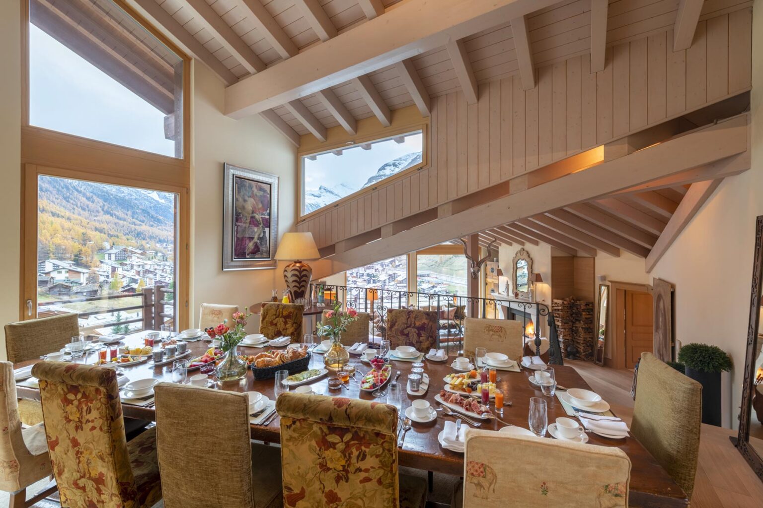 Luxurious dining room with a large wooden table set for breakfast, featuring an array of gourmet dishes. The space is illuminated by natural light from large windows, showcasing a scenic mountain view. Elegant decor includes plush chairs and stylish wood beams, creating a warm and inviting atmosphere. | Only on SopranoVillas