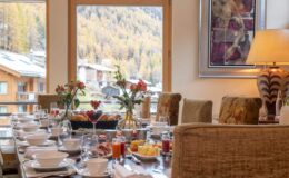 Elegant dining room set for breakfast, featuring a long table with white dishware, fresh flowers, and a view of autumn foliage outside the window. Cozy atmosphere with warm lighting and tasteful decor.