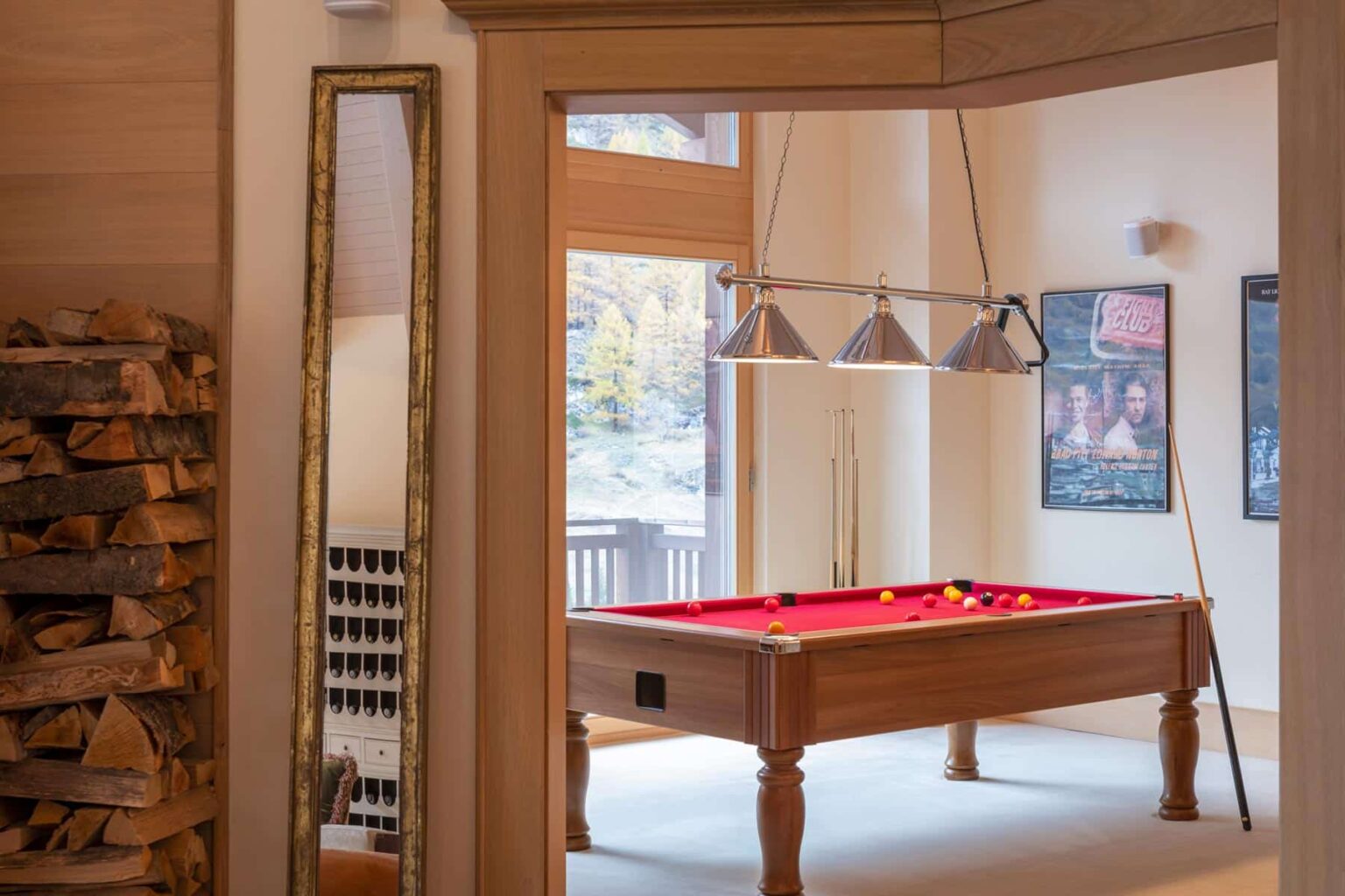 Interior view of a game room featuring a red felt pool table, overhead lighting, and a wooden mirror. The space includes a stack of firewood and large windows that provide natural light, creating a cozy atmosphere. | Only on SopranoVillas