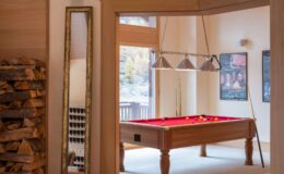 Interior view of a game room featuring a red felt pool table, overhead lighting, and a wooden mirror. The space includes a stack of firewood and large windows that provide natural light, creating a cozy atmosphere.