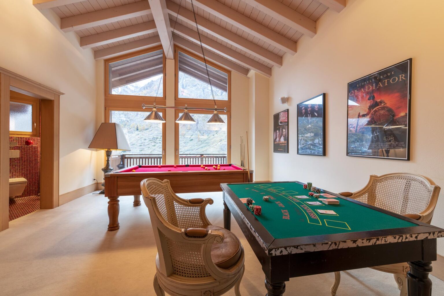 Luxurious game room featuring a pool table and a green felt poker table, with large windows offering scenic mountain views and rustic wooden beams overhead. | Only on SopranoVillas