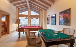 Luxurious game room featuring a pool table and a green felt poker table, with large windows offering scenic mountain views and rustic wooden beams overhead.