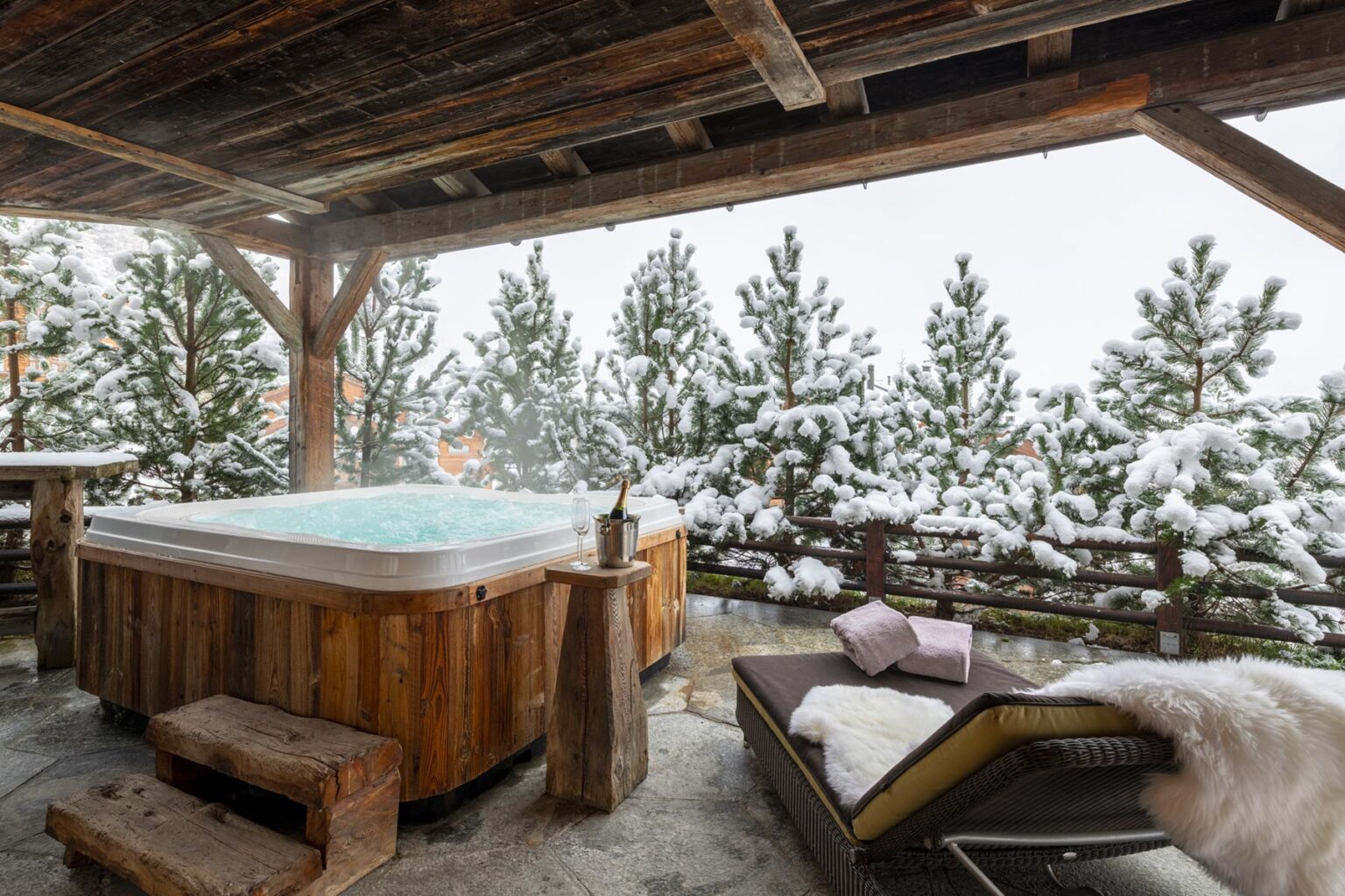 Cozy outdoor hot tub surrounded by snow-covered trees, with a wooden deck and lounge chair, ideal for a winter relaxation retreat. | Only on SopranoVillas