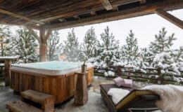 Cozy outdoor hot tub surrounded by snow-covered trees, with a wooden deck and lounge chair, ideal for a winter relaxation retreat.