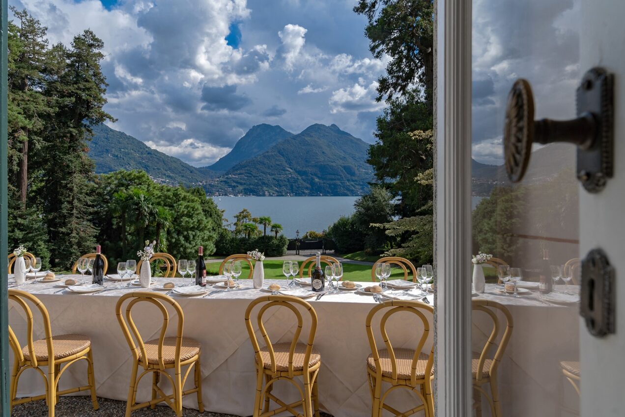 Scenic view of a dining setup with elegant chairs, overlooking a lake and mountains under a partly cloudy sky, framed by an open door. Ideal for outdoor events or weddings. | Only on SopranoVillas