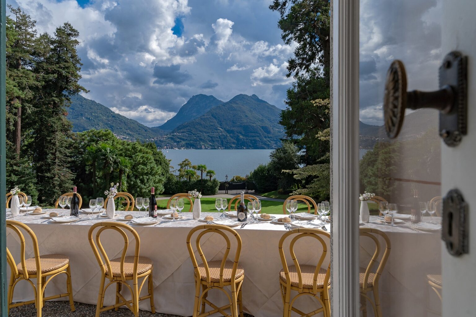 Scenic view of a dining setup with elegant chairs, overlooking a lake and mountains under a partly cloudy sky, framed by an open door. Ideal for outdoor events or weddings. | Only on SopranoVillas