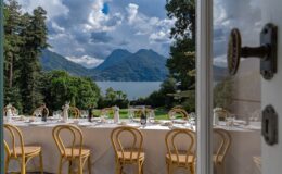 Scenic view of a dining setup with elegant chairs, overlooking a lake and mountains under a partly cloudy sky, framed by an open door. Ideal for outdoor events or weddings.