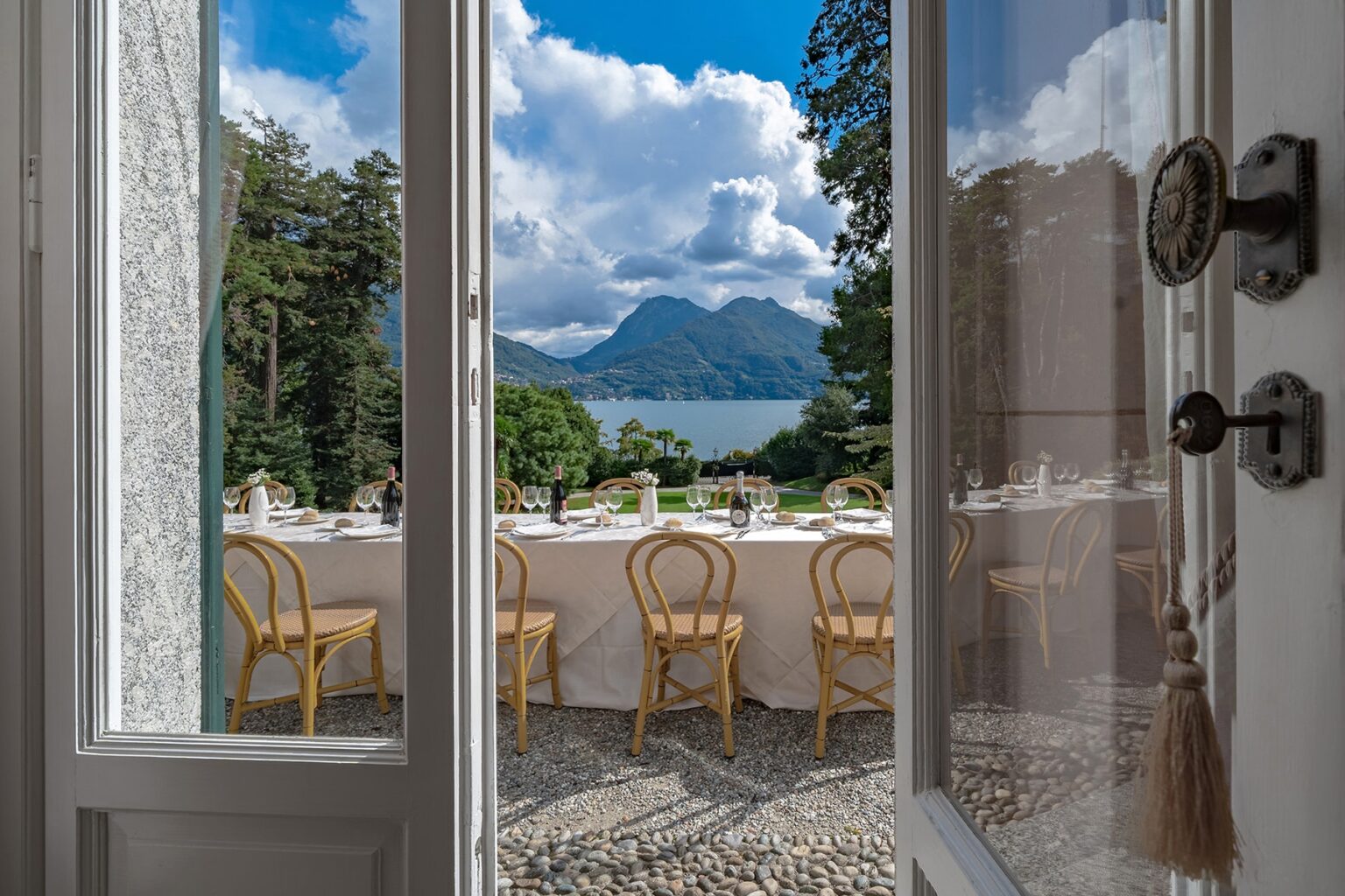 View from an open door showcasing a beautifully set outdoor dining table with wicker chairs, overlooking a serene lake and mountains under a partly cloudy sky. Perfect for event planning and scenic dining experiences. | Only on SopranoVillas