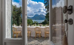 View from an open door showcasing a beautifully set outdoor dining table with wicker chairs, overlooking a serene lake and mountains under a partly cloudy sky. Perfect for event planning and scenic dining experiences.