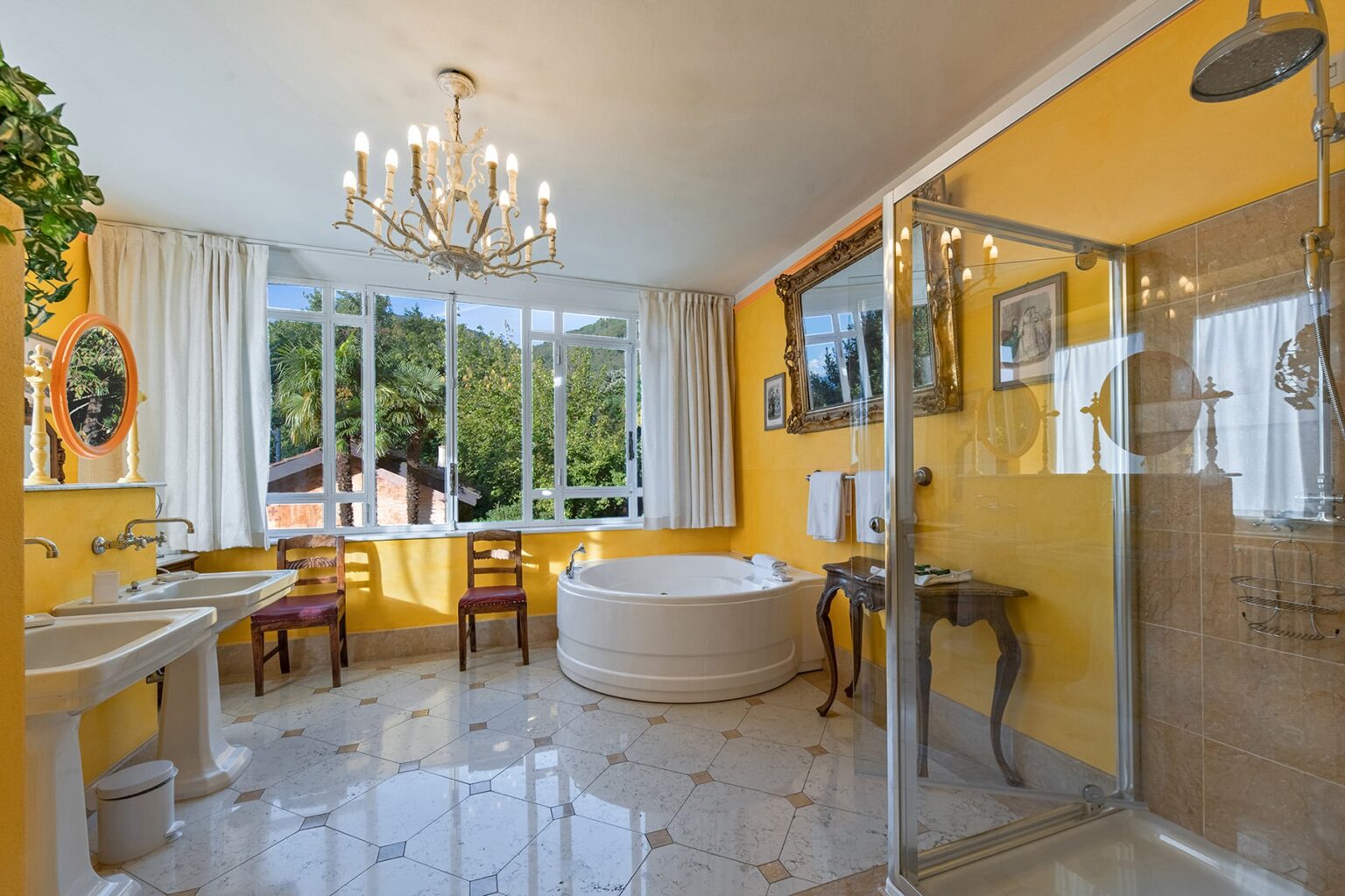 Bright and modern bathroom featuring a yellow color scheme, a large freestanding bathtub, a glass shower, elegant chandelier, and stylish furnishings, set against a backdrop of large windows offering natural light and a view of greenery. | Only on SopranoVillas