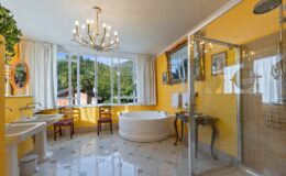 Bright and modern bathroom featuring a yellow color scheme, a large freestanding bathtub, a glass shower, elegant chandelier, and stylish furnishings, set against a backdrop of large windows offering natural light and a view of greenery.