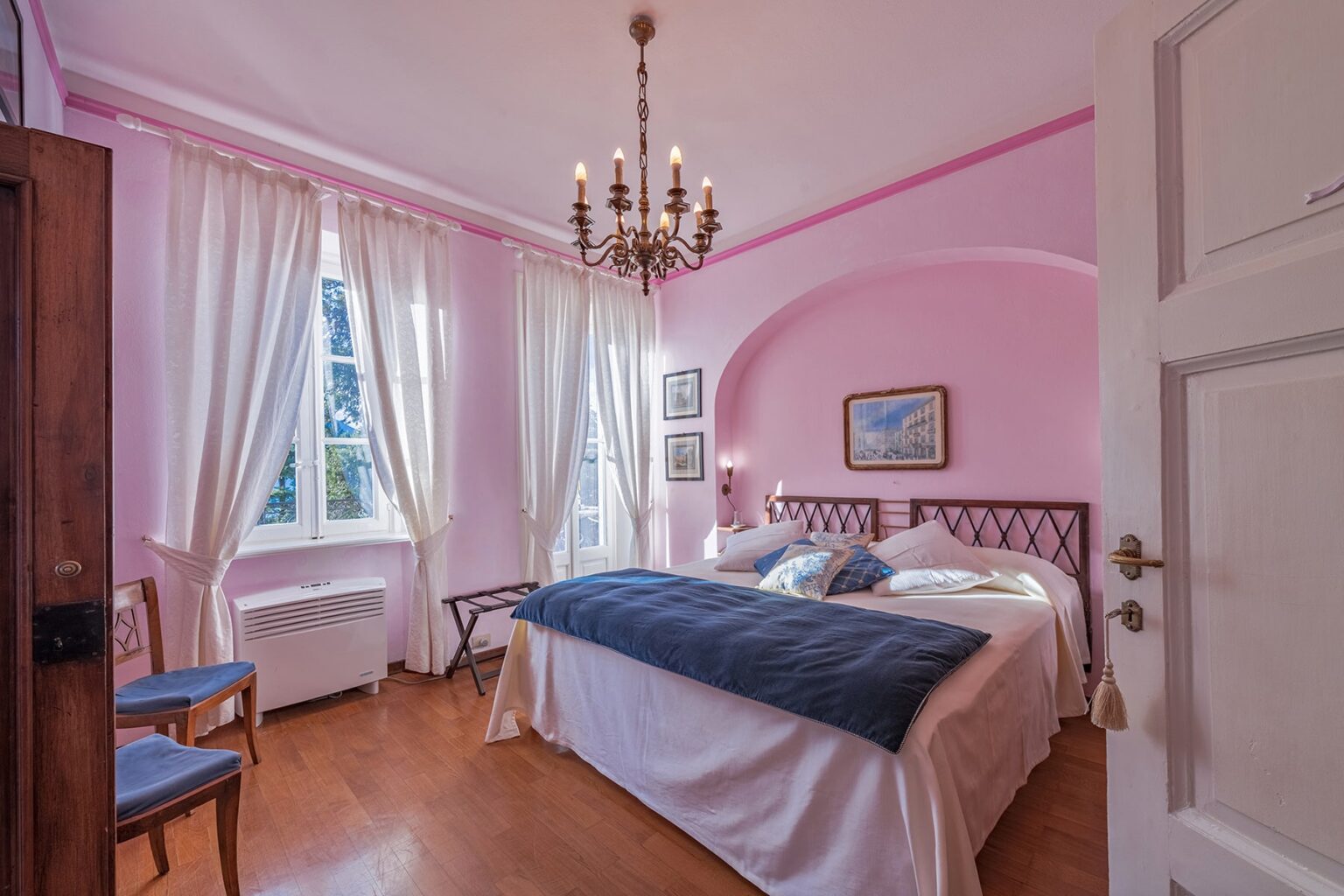 Cozy bedroom featuring soft pink walls, elegant chandelier, large windows with sheer curtains, and a comfortable double bed with a blue throw blanket. The room is adorned with artwork and has wooden flooring, creating a warm and inviting atmosphere. | Only on SopranoVillas