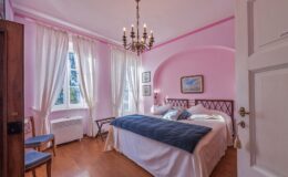 Cozy bedroom featuring soft pink walls, elegant chandelier, large windows with sheer curtains, and a comfortable double bed with a blue throw blanket. The room is adorned with artwork and has wooden flooring, creating a warm and inviting atmosphere.