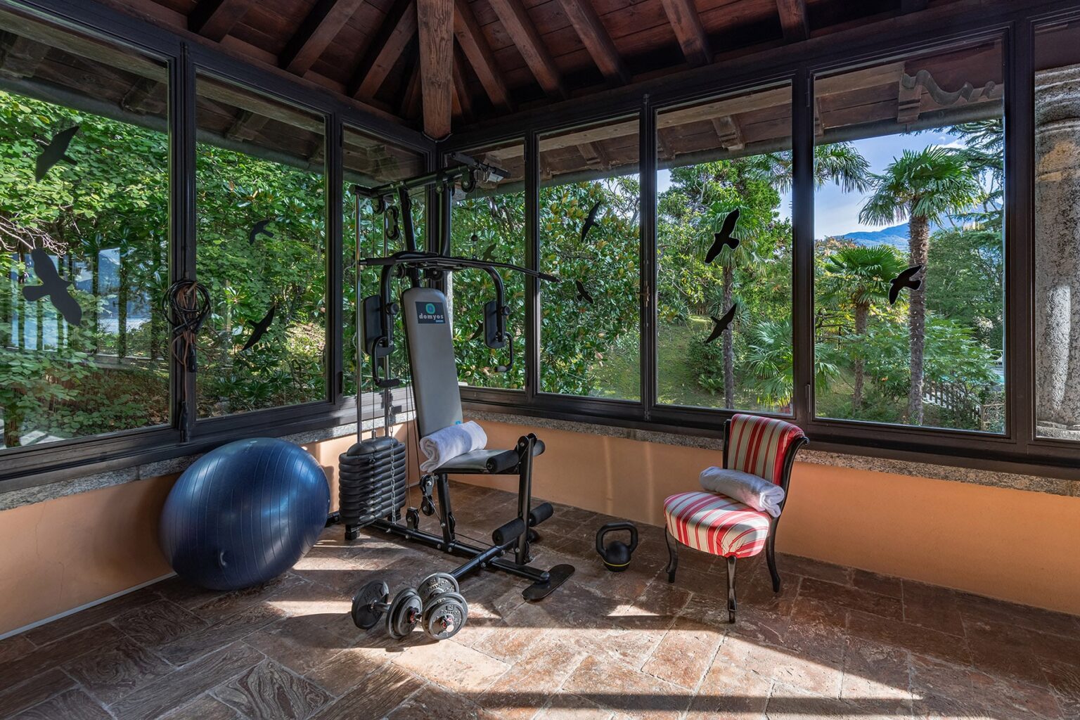 Home gym with exercise equipment, including a weight bench and stability ball, surrounded by large windows offering a view of greenery. The space features a rustic ceiling and a colorful chair, creating a bright and inviting workout environment. | Only on SopranoVillas