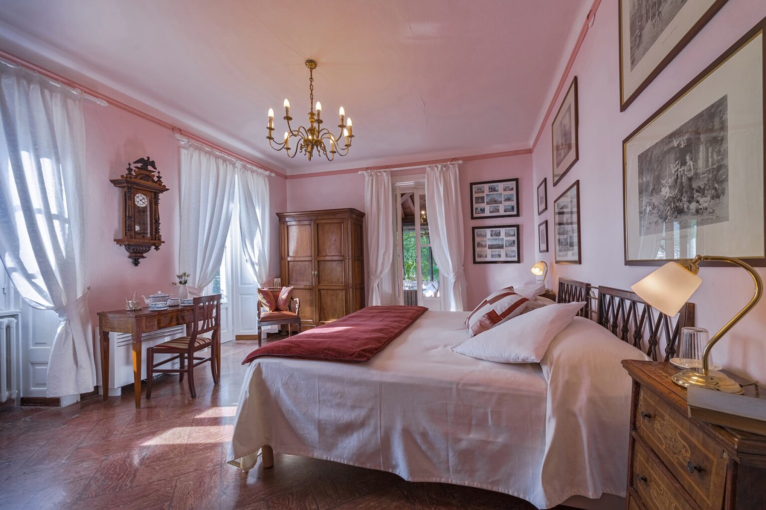 Cozy and elegant bedroom featuring pink walls, a chandelier, and vintage decor. The room includes a large bed with white linens, a wooden nightstand with a lamp, and a desk with a chair. Natural light streams through the curtains, enhancing the inviting atmosphere. | Only on SopranoVillas