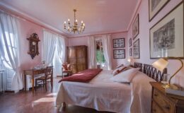 Cozy and elegant bedroom featuring pink walls, a chandelier, and vintage decor. The room includes a large bed with white linens, a wooden nightstand with a lamp, and a desk with a chair. Natural light streams through the curtains, enhancing the inviting atmosphere.