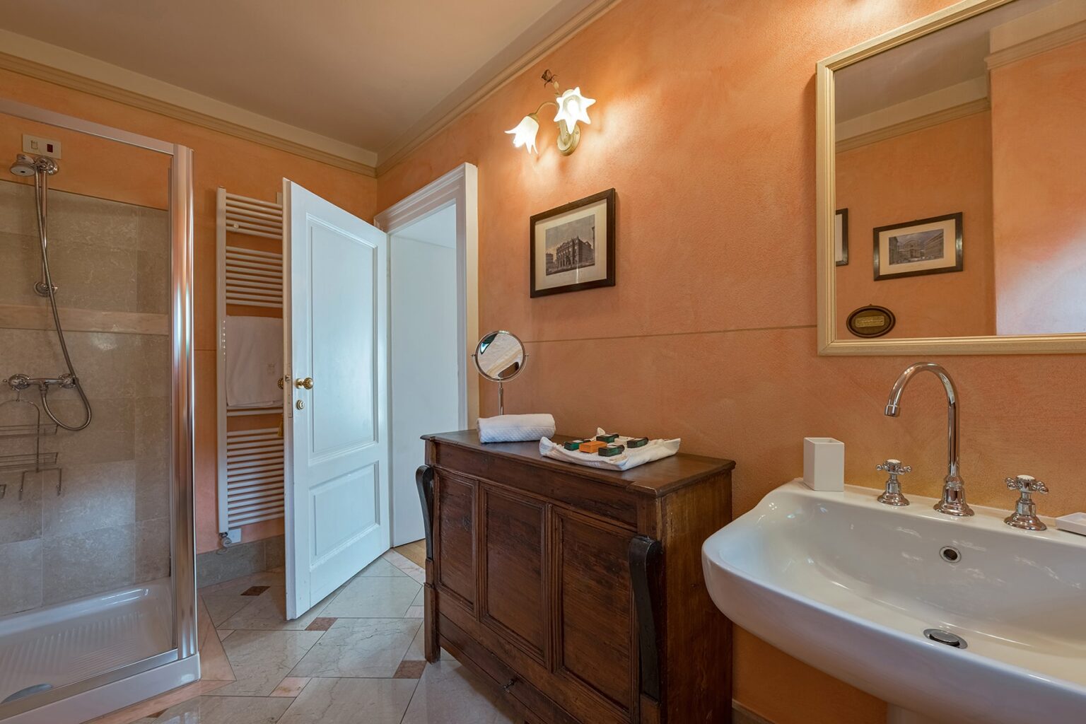 Modern bathroom featuring an orange accent wall, a spacious shower, a wooden vanity with decorative items, and a large mirror, providing a warm and inviting atmosphere. | Only on SopranoVillas