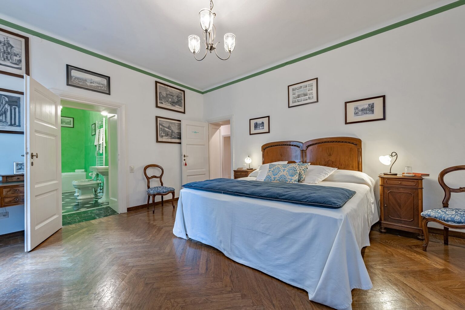 Elegant bedroom with a large double bed, wooden furniture, and vintage decor, featuring an en-suite bathroom visible through an open door. The room is well-lit with a chandelier and has a warm, inviting atmosphere. | Only on SopranoVillas