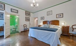 Elegant bedroom with a large double bed, wooden furniture, and vintage decor, featuring an en-suite bathroom visible through an open door. The room is well-lit with a chandelier and has a warm, inviting atmosphere.