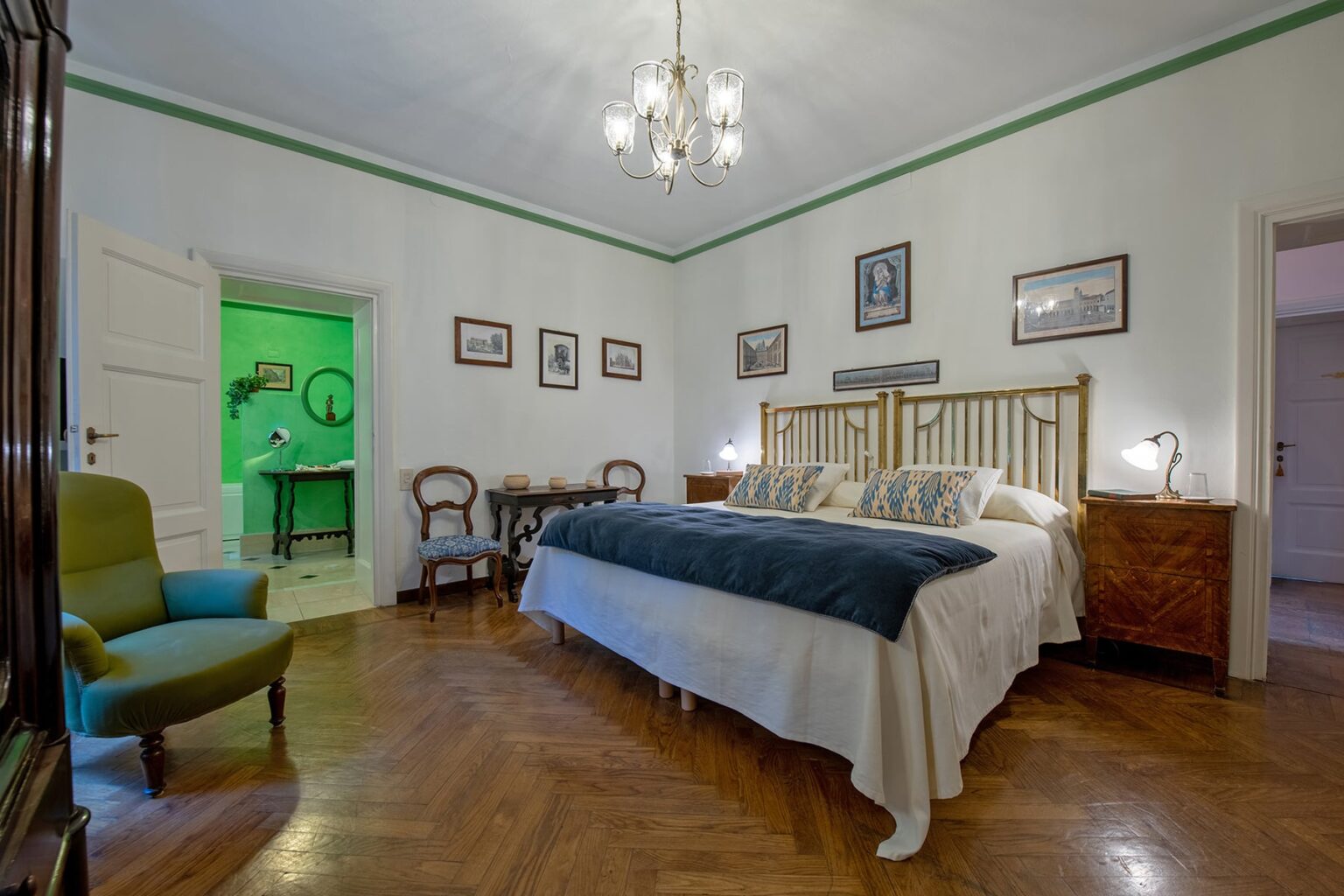Spacious and elegantly decorated bedroom featuring a large bed with plush bedding, a vintage armchair, and wooden furniture. The room is illuminated by a stylish chandelier and showcases framed artwork on the walls, creating a cozy and inviting atmosphere. | Only on SopranoVillas
