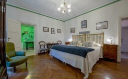 Spacious and elegantly decorated bedroom featuring a large bed with plush bedding, a vintage armchair, and wooden furniture. The room is illuminated by a stylish chandelier and showcases framed artwork on the walls, creating a cozy and inviting atmosphere.