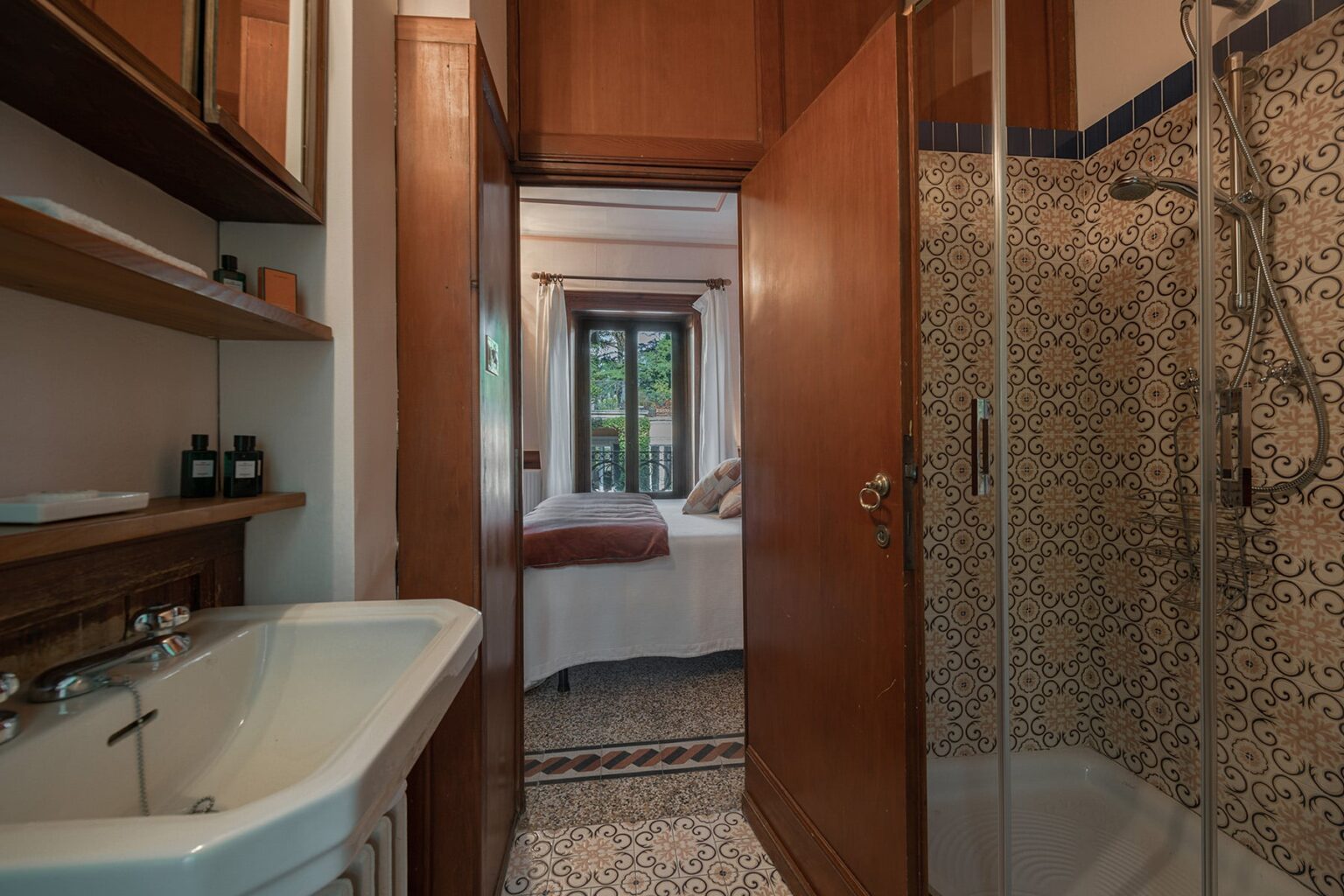 Modern bathroom with a glass shower, sink, and wooden cabinetry, leading to a cozy bedroom with large windows. Decorative tiled walls add a touch of elegance. | Only on SopranoVillas