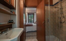 Modern bathroom with a glass shower, sink, and wooden cabinetry, leading to a cozy bedroom with large windows. Decorative tiled walls add a touch of elegance.