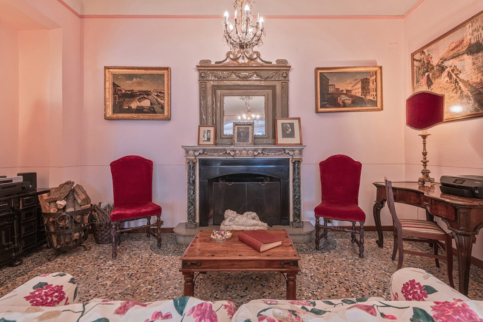 Cozy vintage living room featuring ornate decor, two red upholstered chairs, a marble fireplace with a decorative mirror, and classic artwork on the walls. The room is illuminated by a chandelier, creating an inviting atmosphere. | Only on SopranoVillas
