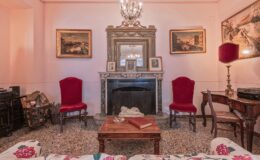 Cozy vintage living room featuring ornate decor, two red upholstered chairs, a marble fireplace with a decorative mirror, and classic artwork on the walls. The room is illuminated by a chandelier, creating an inviting atmosphere.