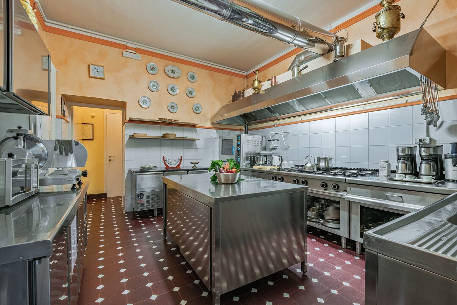 Modern commercial kitchen with stainless steel equipment, spacious island, and decorative plates on the wall, showcasing a clean and organized cooking environment. | Only on SopranoVillas